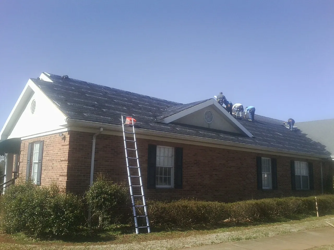 Crew tearing shingles off a brick ranch roof with a ladder set — Emergency Roof Repair work by our crew in Northbridge