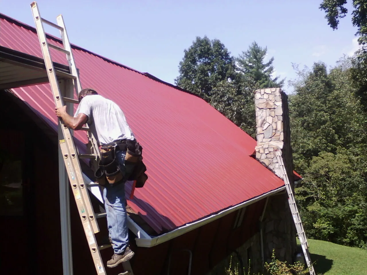 Roofer on a ladder beside a red metal roof and stone chimney — Soffit & Fascia Repair by Roofing Brothers in Northbridge
